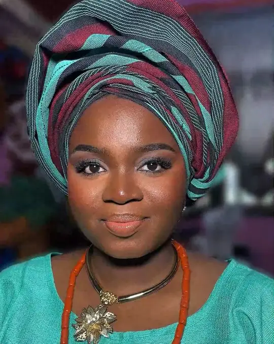 A smiling woman in a teal gele and coral beads
